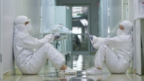 two tired doctors in protective wear sitting on floor in hallway