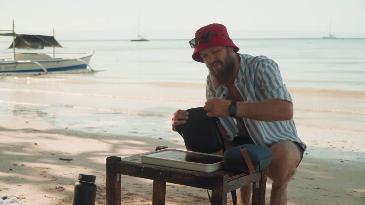 Uomo che prepara il tè su tavolo da spiaggia, cappello rosso, barca sullo sfondo, intima cerimonia del tè in riva al mare al tramonto con luce calda, onde leggere, movimenti rituali, bollitore artigianale, tazza, rilassamento consapevole