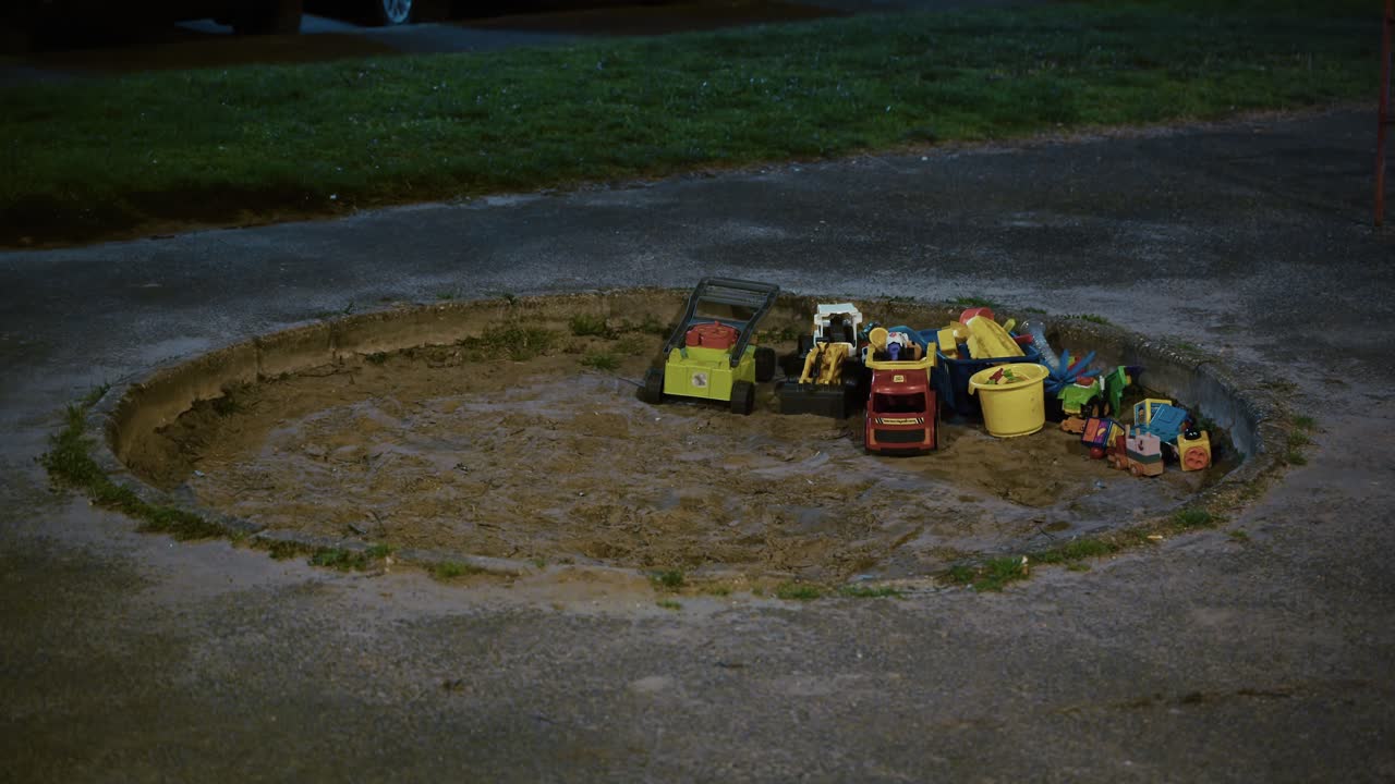 Plastic toy trucks and buckets left in a round sandbox at night under dim streetlight
