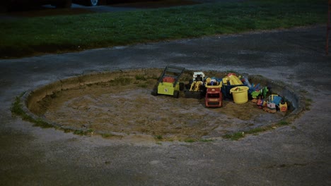 plastic toy trucks and buckets left in a round sandbox at night under dim streetlight