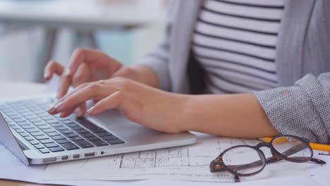 Close-Up-Of-Female-Architect-In-Office-At-Desk-Working-On-Plans-For-New-Building-Using-Laptop