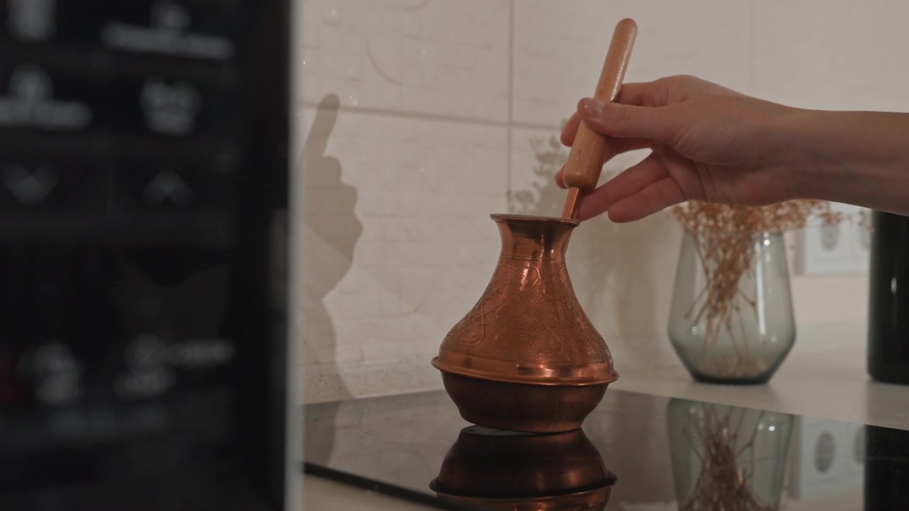 Un cezve en cuivre sur la cuisinière, une main remuant le café, un rituel pour amateur de café. Moment intime avec une plaque de cuisson sombre et réfléchissante, des fleurs séchées dans un vase en verre et une douce lumière ambiante. Gros plan sur l'infusion tactile, grain cinématographique, dans une cuisine douillette.