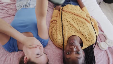 Happy-diverse-teenage-female-friends-lying-on-their-backs-on-bed-using-smartphone-and-taking-selfie