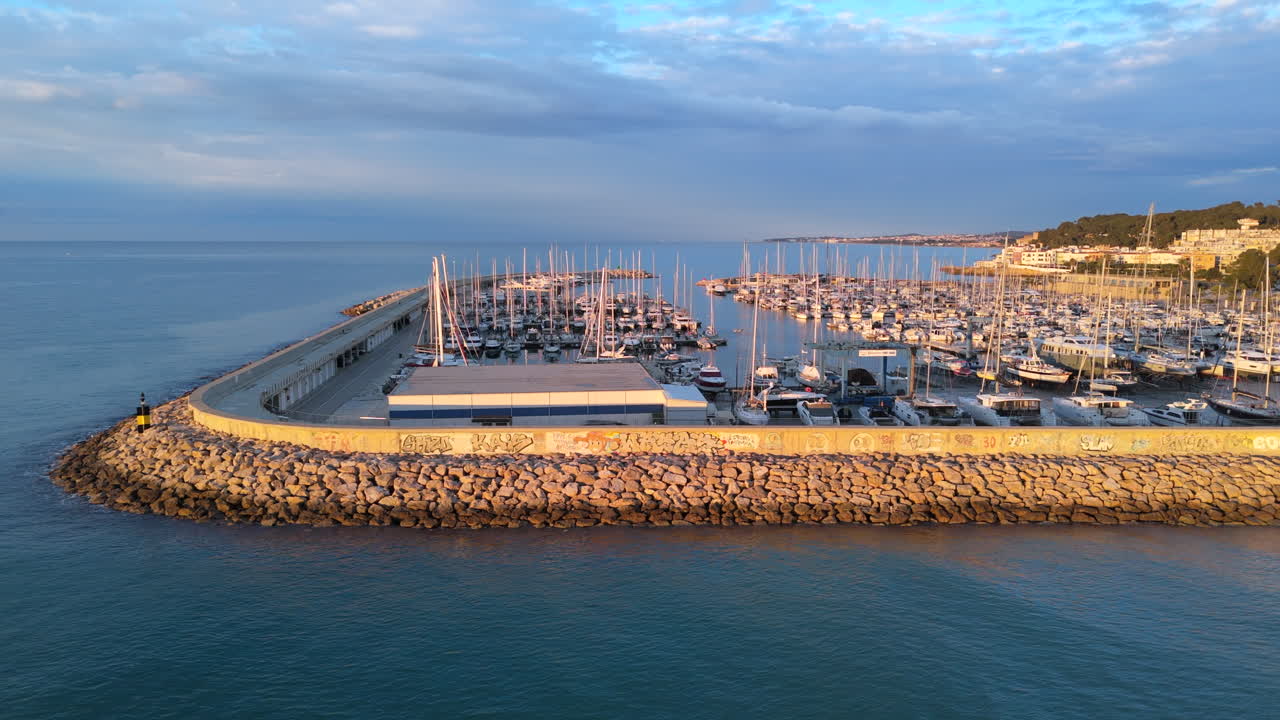 A slow, cinematic aerial traveling along a marina’s breakwater at sunrise. The golden light enhances the yachts, calm sea, and coastal beauty. Perfect video for travel, tourism, and maritime projects