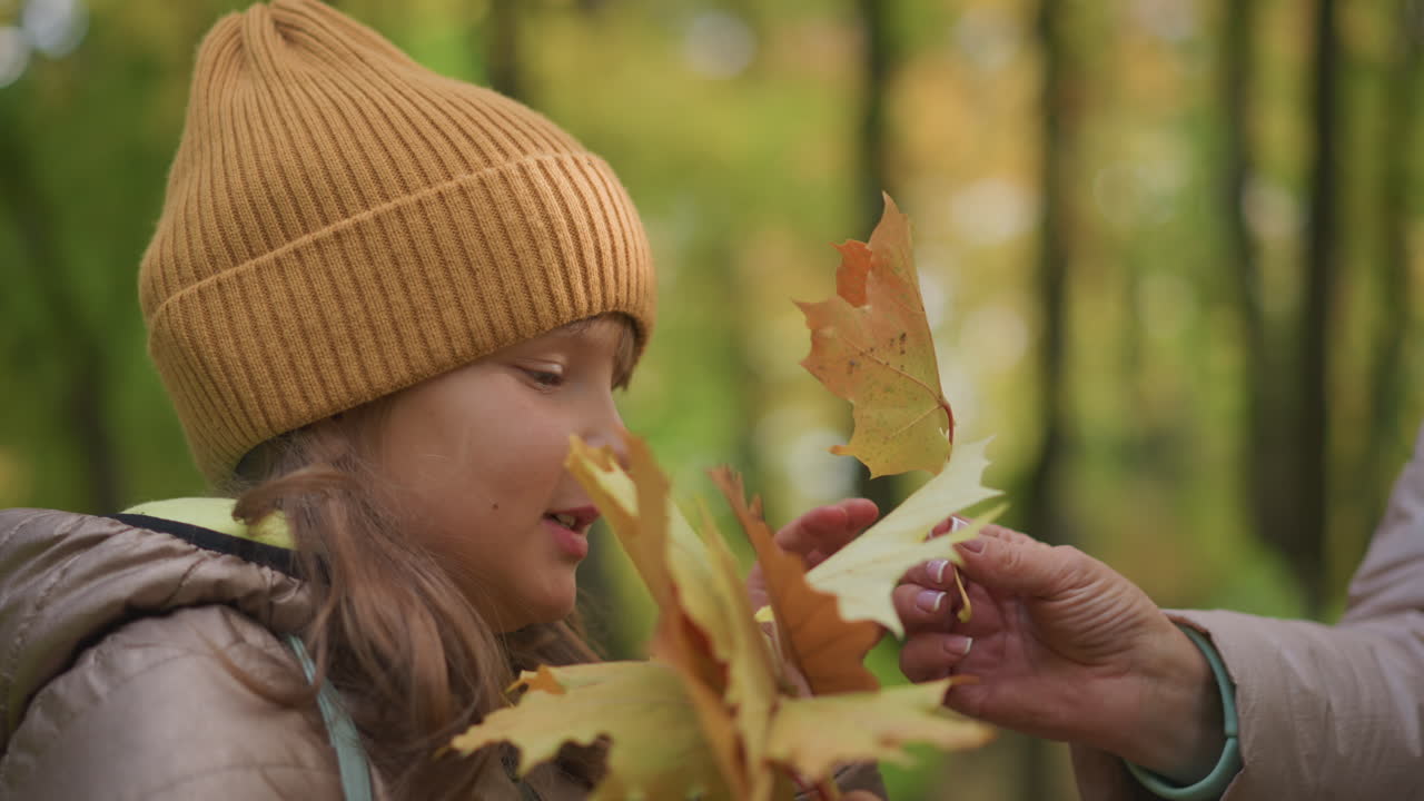 side profile of young girl in brown jacket and mustard hat gently smelling autumn leaves with eyes closed, standing in peaceful forest as unseen person offers her another leaf