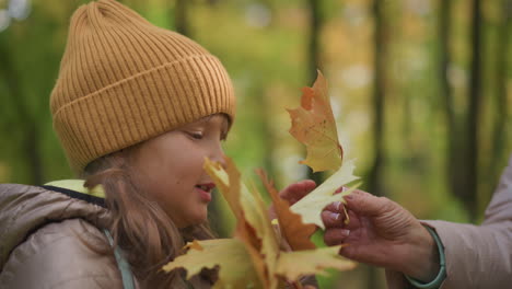 side profile of young girl in brown jacket and mustard hat gently smelling autumn leaves with eyes closed, standing in peaceful forest as unseen person offers her another leaf