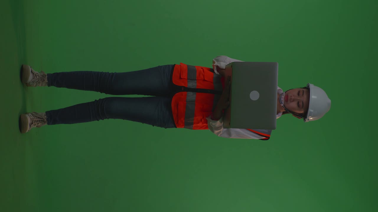 cuerpo lleno de ingeniera asiática con casco de seguridad trabajando en una computadora portátil mientras está de pie en el estudio de fondo de pantalla verde, durante la puesta o el amanecer