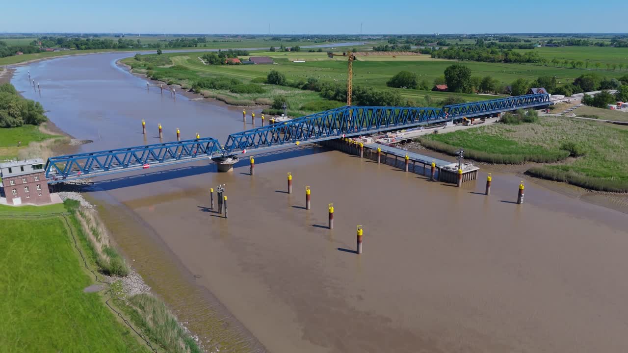Low angle drone shot of the closed Friesenbrücke railway bridge in Northern Germany. Shows the full span, construction site, and surrounding river landscape on a clear summer day.