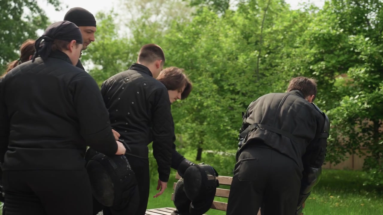Gruppo di persone in equipaggiamento protettivo nero che si prepara per un'attività all'aperto, tenendo in mano caschi e indossando tute imbottite. Scena ambientata su un sentiero circondato da alberi verdi con luce diurna intensa.