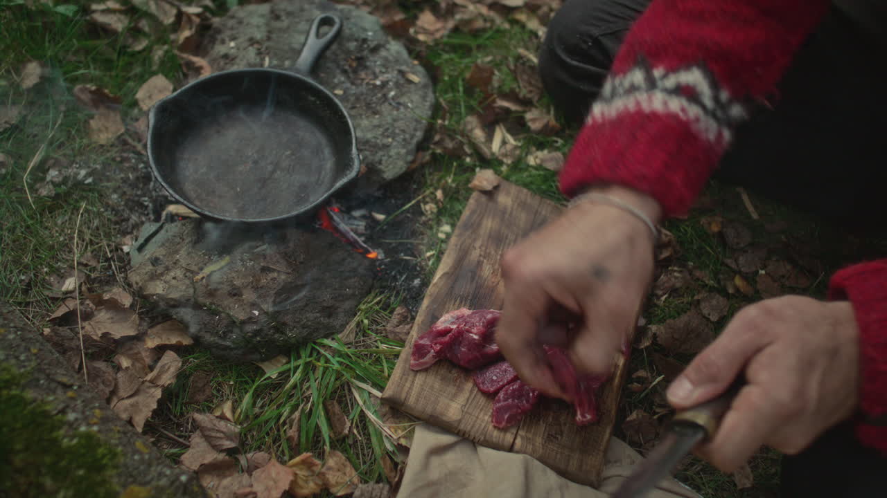 Hands of Man Cutting Raw Meat and Placing in Skillet over Campfire in Forest