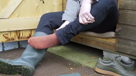 man sitting at garden shed taking off green wellington boots after mowing the lawn