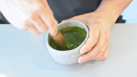 Handheld-motion-close-up-shot-of-a-pair-of-hands-whisking-and-mixing-match-green-tea-in-the-handmade-ceramic-bowl-with-bamboo-whisk,-preparation-of-traditional-Japanese-beverages,-specialty-drinks