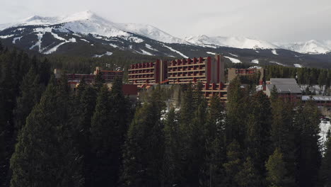 Rising-drone-footage,-over-pine-trees,-of-the-Beaver-Run-Resort-and-Conference-Center,-Breckenridge-mountain-range-,-and-a-ski-lift-with-skiers-and-snowboarders-in-Summit-County-Colorado