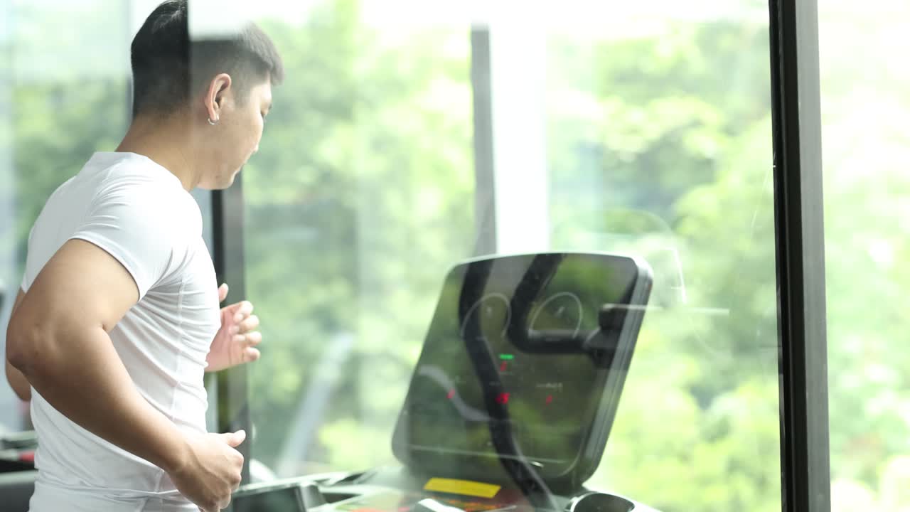 A man exercises on a treadmill in a well-lit gym with large windows, showcasing determination and fitness
