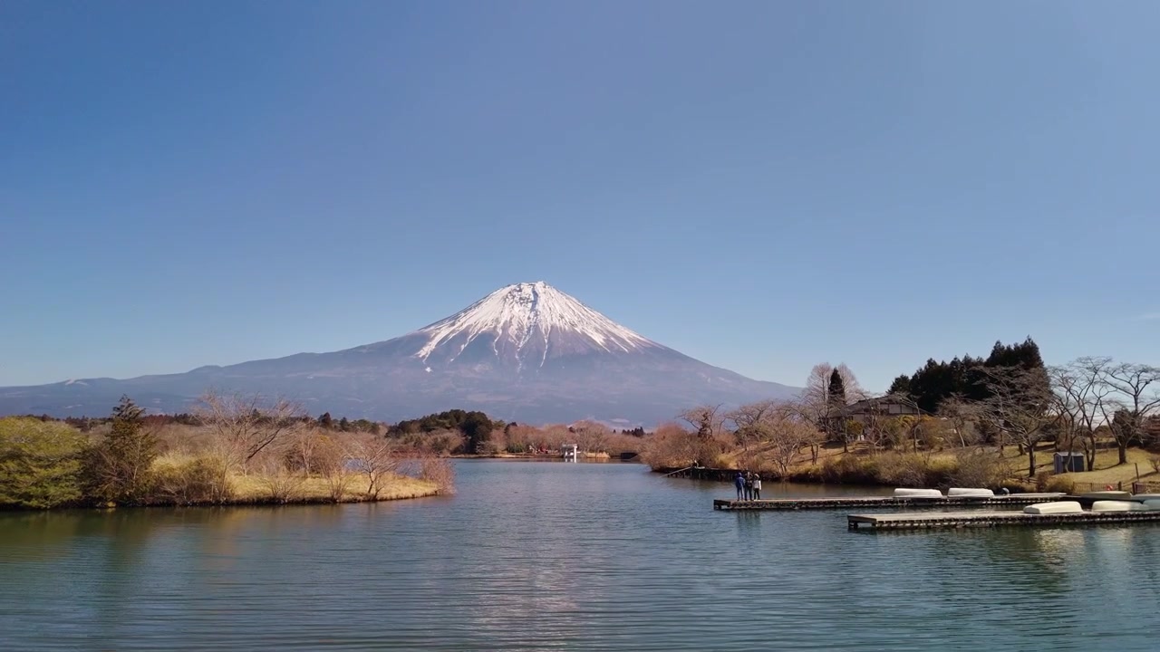 一幅風景優美的廣角照片，展示了富士山在寧靜湖泊上方的壯麗景色，遊客們站在木製碼頭上欣賞美景。