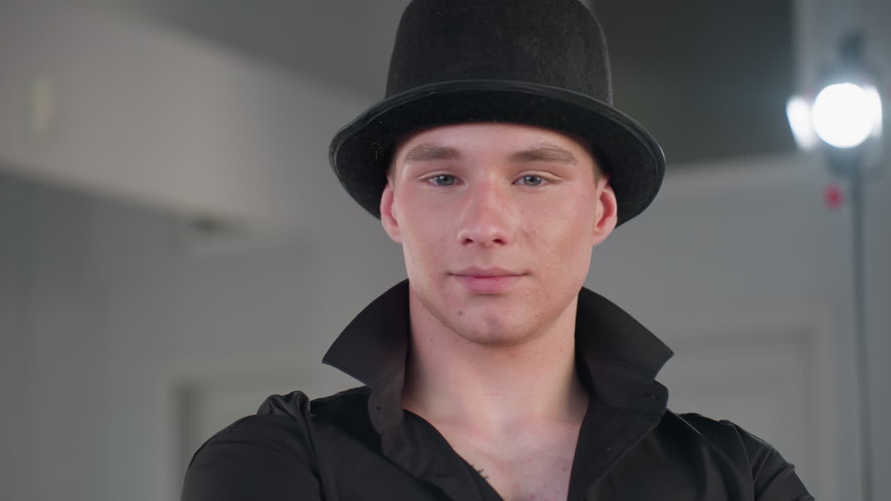 handsome young man in black shirt and top hat turns and smiles toward camera inside minimal studio set with soft light stand in background exuding charming confidence and stylish attitude