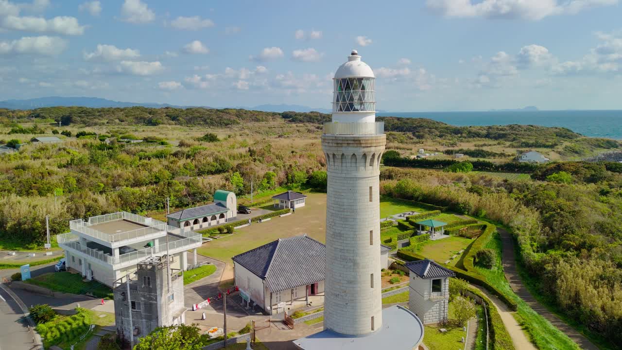 An aerial shot circles the historic Izumo Hinomisaki Lighthouse on a sunny day, showcasing the tallest stone lighthouse in Japan against a backdrop of green hills and the sea
