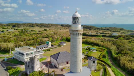 an aerial shot circles the historic izumo hinomisaki lighthouse on a sunny day, showcasing the tallest stone lighthouse in japan against a backdrop of green hills and the sea