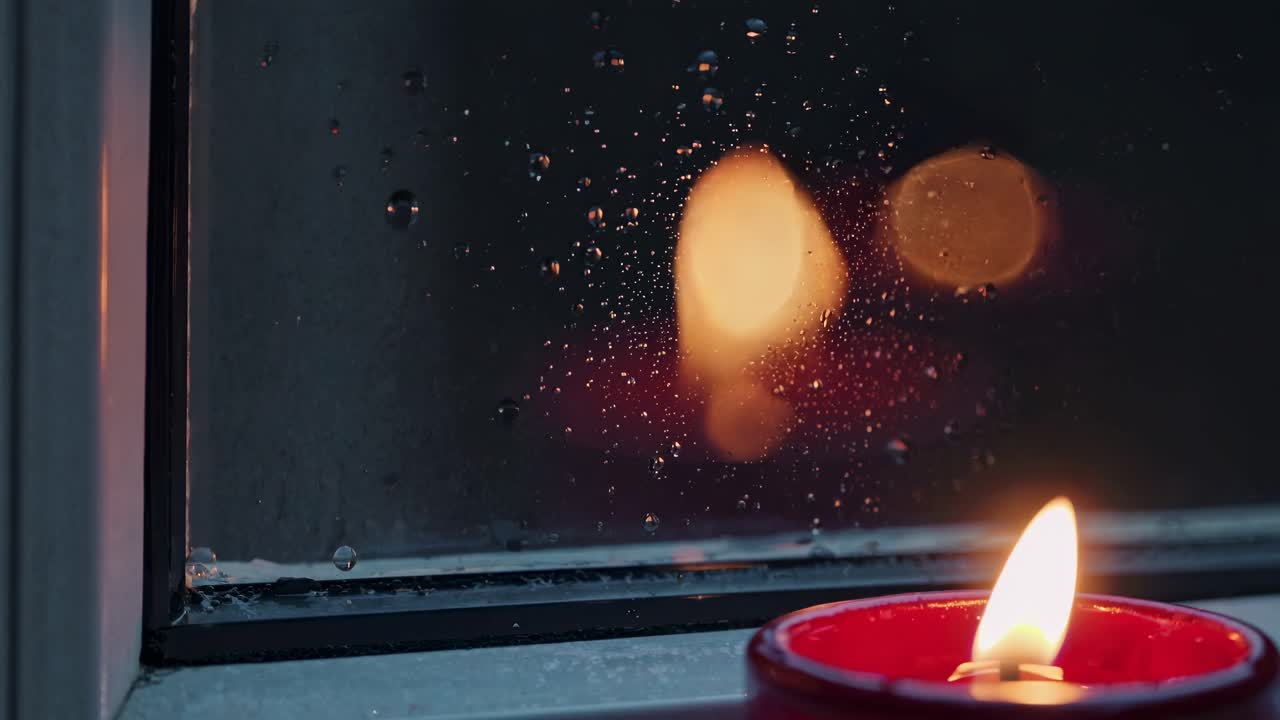 Cozy video scene with a close-up angle of a candle by a rainy window, capturing warm light