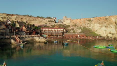 Popeye-Village-in-Anchor-Bay,-Gozo-Island,-Malta-wooden-Village-Houses-by-Water-and-Boats,-Aerial-Drone-Shot-backwards-Dolly