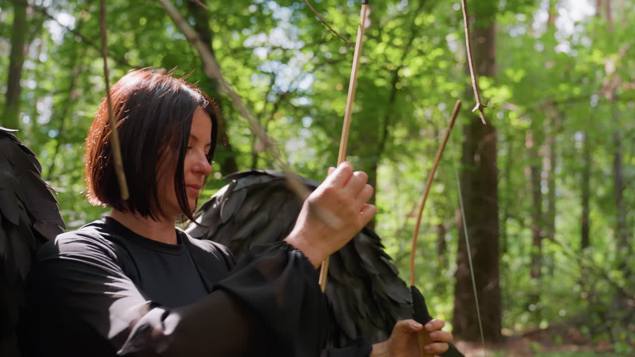 Woman with black wings carefully places arrow on bowstring in green forest, sunlight shining through trees highlighting focus and precision, cinematic moment of concentration