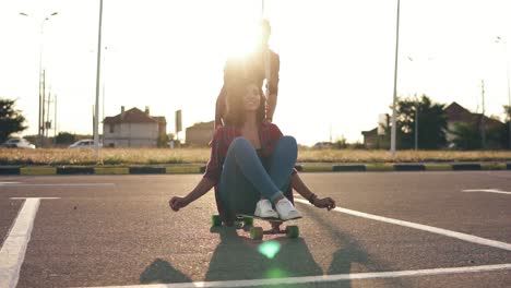 Mujer-Alegre-Sentada-En-Un-Longboard-Y-Levantando-Las-Manos-Alegremente-Mientras-Su-Amiga-La-Empuja-Hacia-Atrás-Durante-La-Puesta-De-Sol