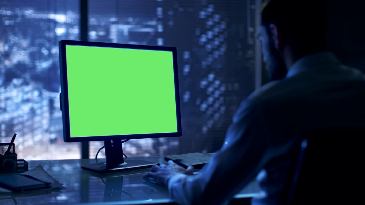 a última hora de la noche el hombre de negocios trabaja en una computadora personal con pantalla verde en su oficina privada con vista a la ventana de la gran ciudad.