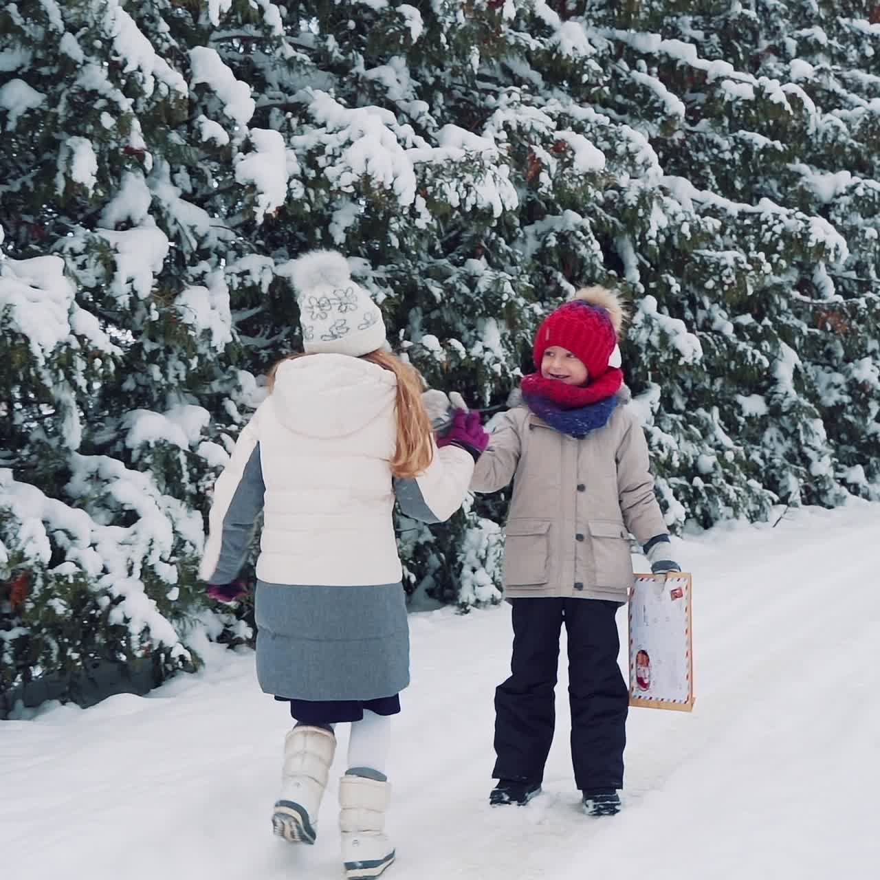 A little boy with an envelope in his hands is waiting for a girl who runs to him on the background of a winter forest on the street. And children are running along a snowy path together on the background of tall fir-trees