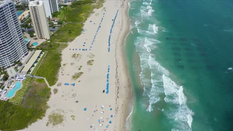 Luftdrohnenaufnahme,-Die-Nach-Vorne-über-Sonnenschirme-Und-Touristen-Am-Strand-Von-Singer-Island-In-Der-Nähe-Des-Ritz-Carlton-Wohngebäudes-Fliegt,-Während-Die-Wellen-Des-Atlantischen-Ozeans-Am-Strand-Krachen