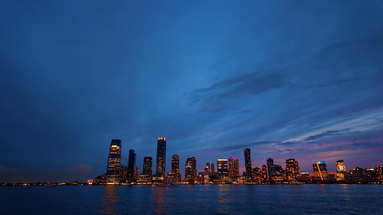 Skyline di Jersey City attraverso il fiume Hudson, New Jersey. Le luci delle torri degli uffici si riflettono sull'acqua increspata sotto nuvole blu al crepuscolo.