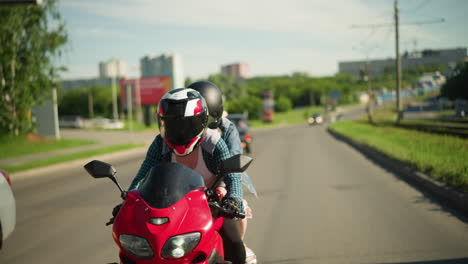 dos hermanas montan una bicicleta eléctrica roja, ambas con cascos, mientras navegan por la carretera de la ciudad, la dama detrás de su tela flota en el viento, edificios, coches y vegetación aparecen en el fondo