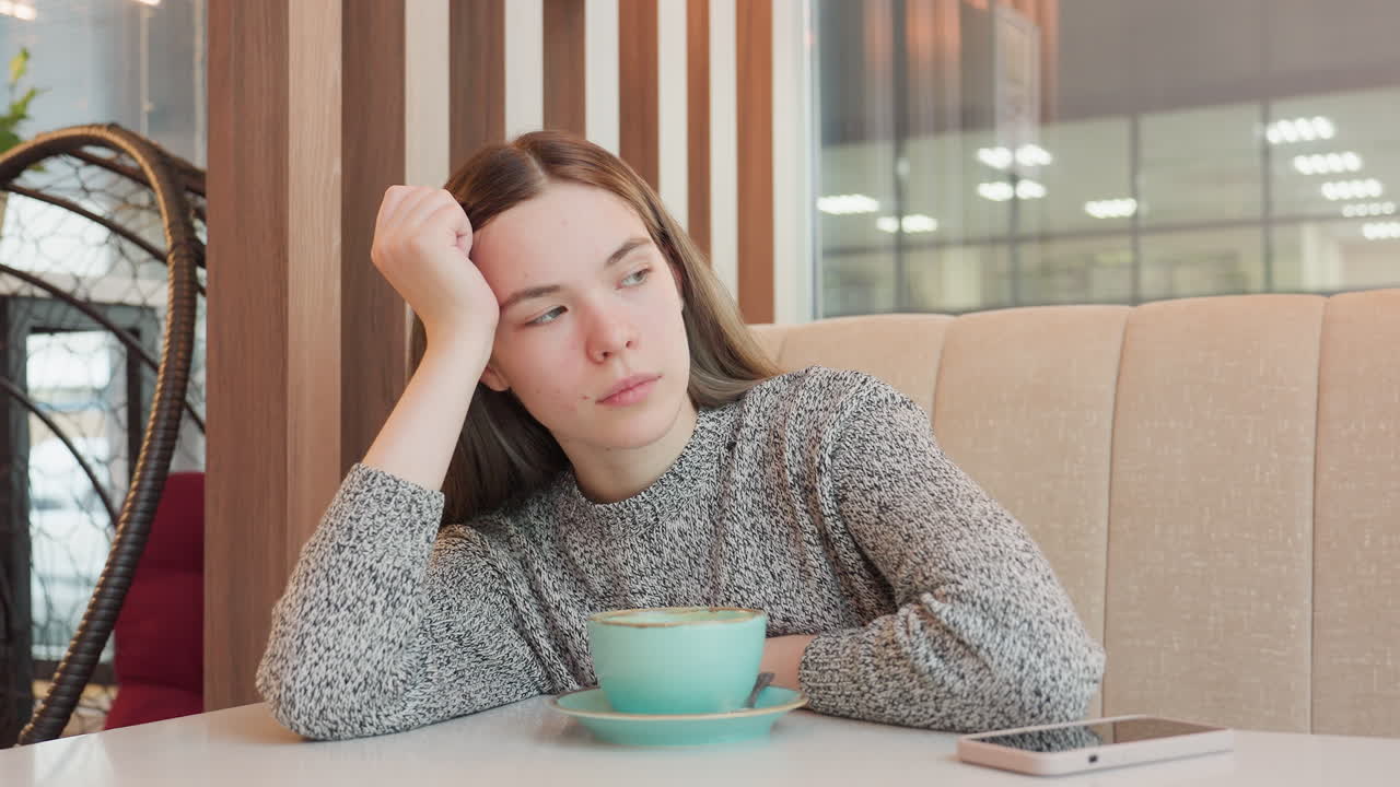 Freelancer gently places one hand on table supporting her head while looking outside attentively with calm thoughtful expression in cozy modern cafe setting beside coffee cup and smartphone