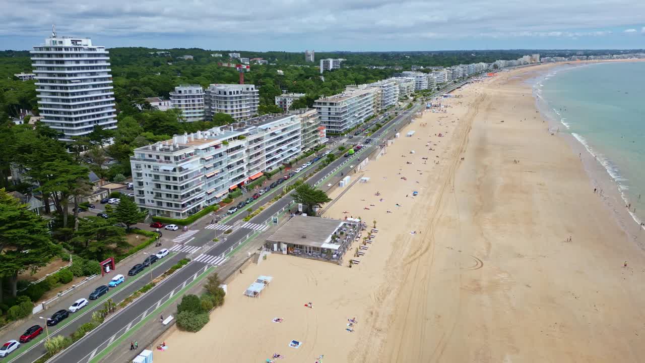 Drone reversing shot of La Baule-Escoublac beach, showing coastal road, buildings, surrounding trees, and beach activity with people walking and sunbathing under umbrellas - France