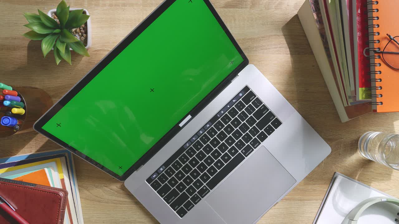 Top down view of a laptop computer with mock up green screen chromakey display on a wooden office desk next to notebook with pens, glasses, and a glass of water. Slow zoom out, close up