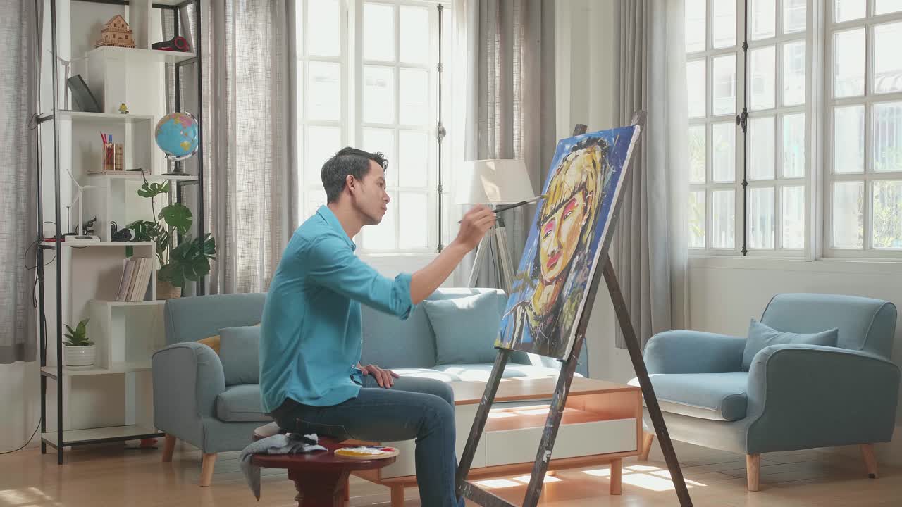 Asian Artist Man Sitting On Round Wooden Chair Without Backrest Holding Paintbrush Mixed Colour And Thinking Before Painting A Girl'S Hair On The Canvas