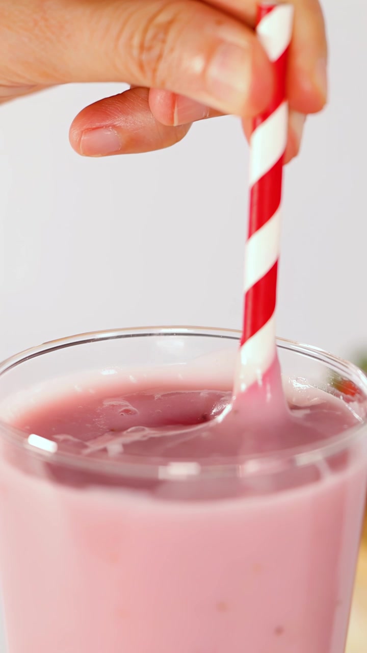 Una mano mescola un frullato di frutta rosa cremoso in un bicchiere usando una cannuccia a strisce rosse, con illuminazione naturale brillante.