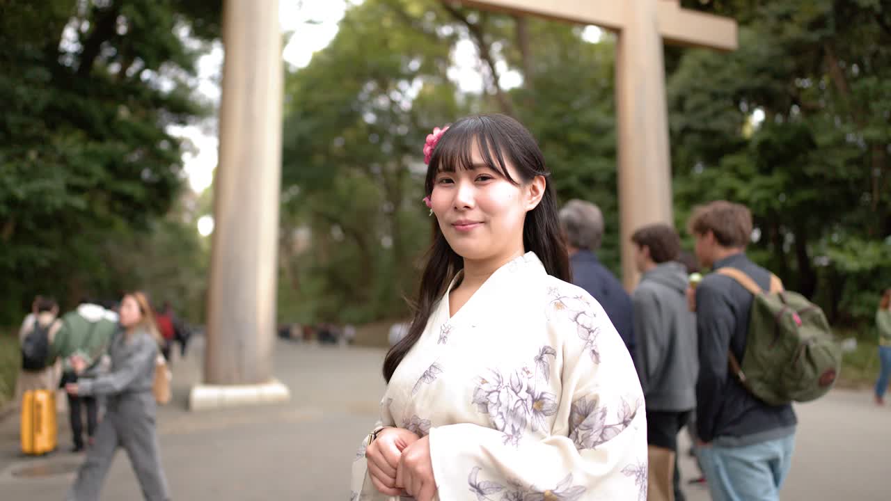 Una mujer japonesa sonriente con un kimono blanco está de pie frente a una gran puerta torii de madera.