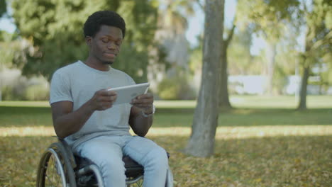 Young Black Man In Wheelchair Spending Time Outdoors With Tablet