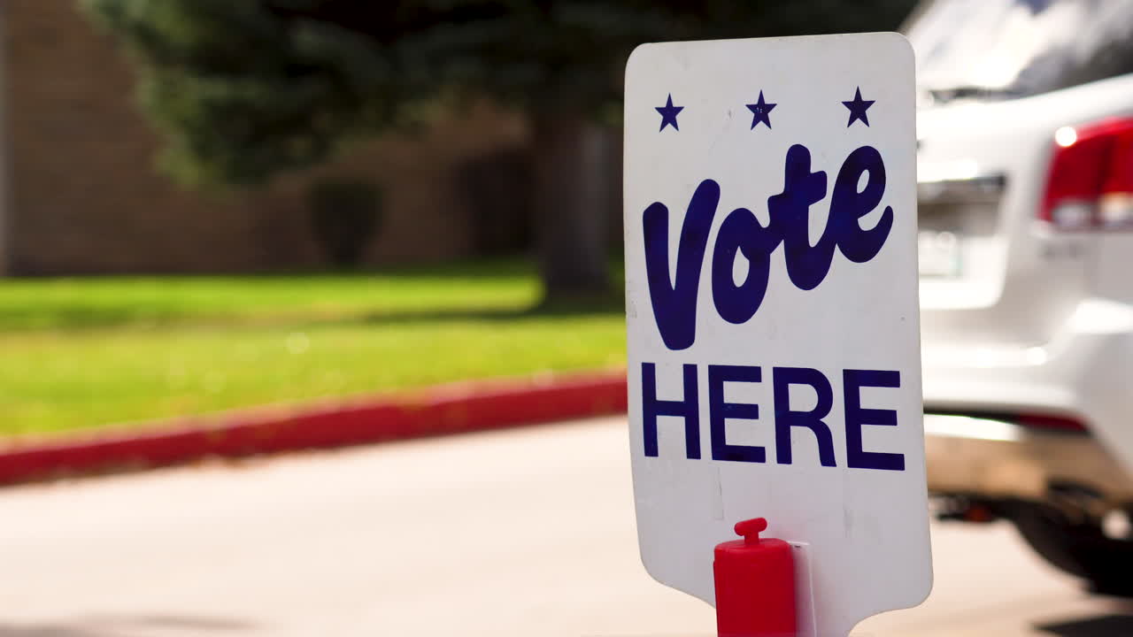 Blue Vote Here Sign Close up with People Driving Cars Walking in the Background