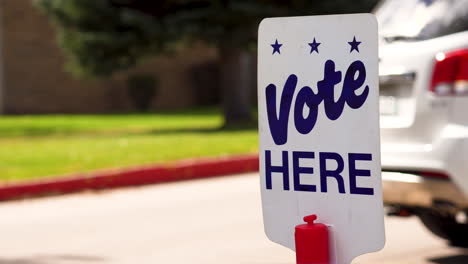 blue vote here sign close up with people driving cars walking in the background