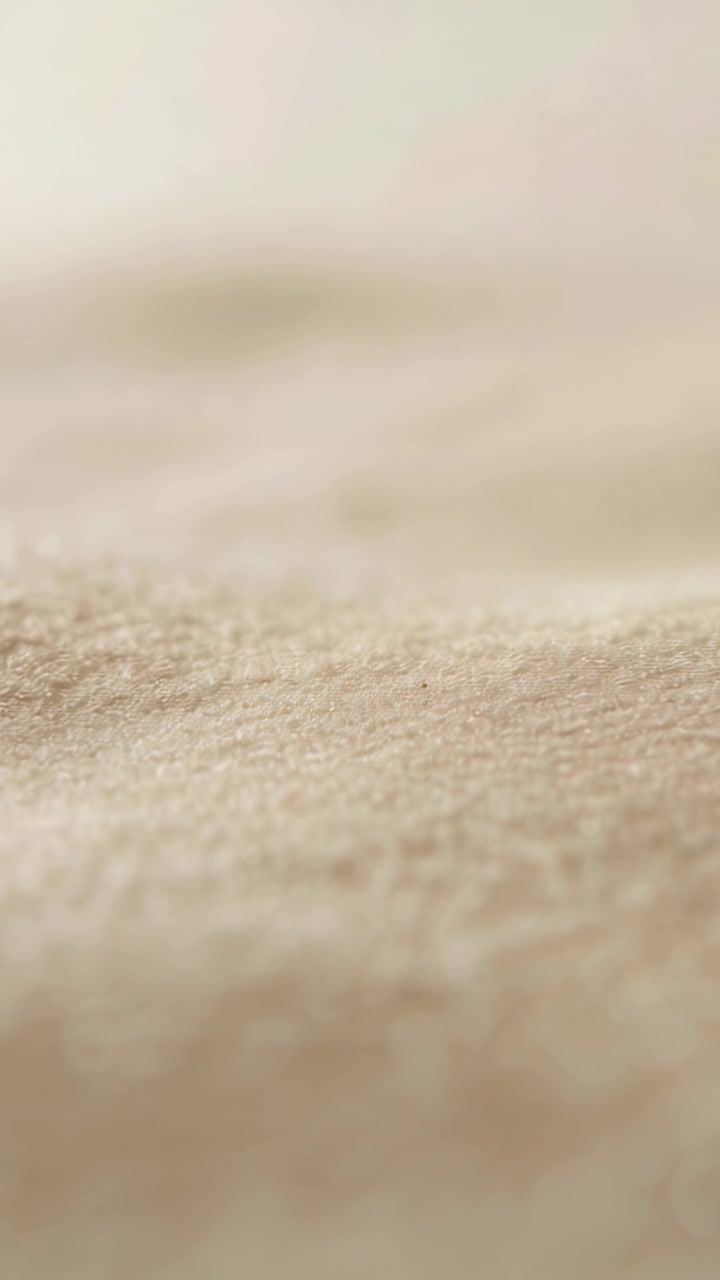 Video verticale: Tessuto beige che scorre rivelando una micro telecamera, con cambio di messa a fuoco e fibre sottili in primo piano.