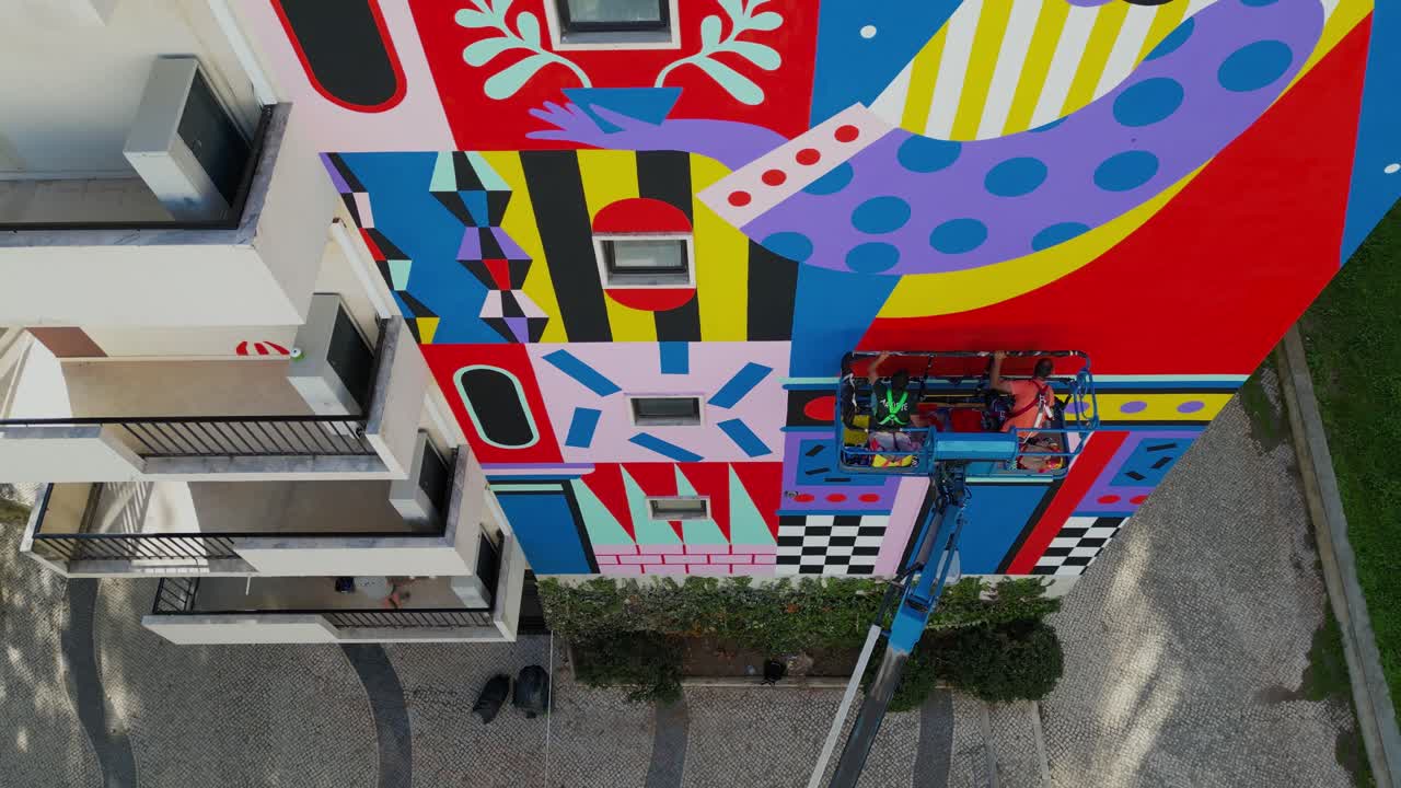 Aerial view of Lisbon: A high-angle photograph captures artists painting a vibrant mural on a building suspended from an elevator, infusing creative energy into the city's urban landscape