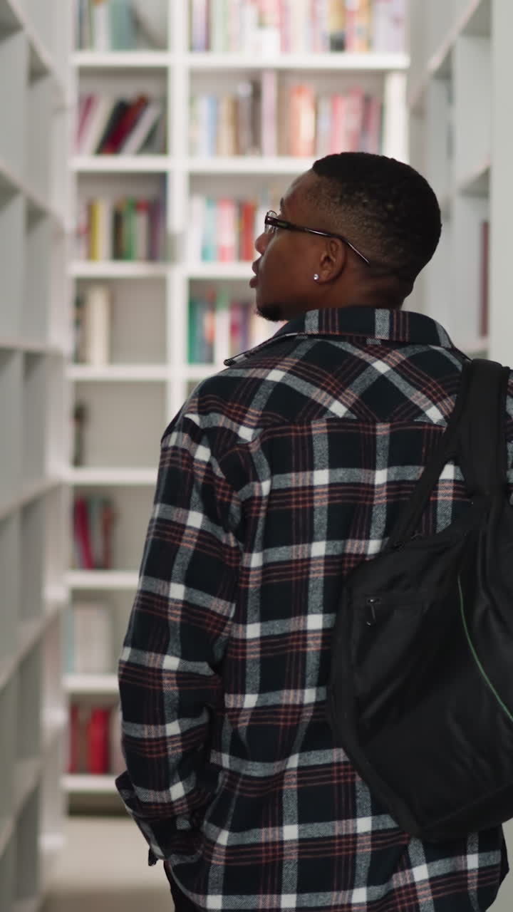 Student walks along aisle in public library. African American man with backpack chooses books on racks in storage. Education and leisure
