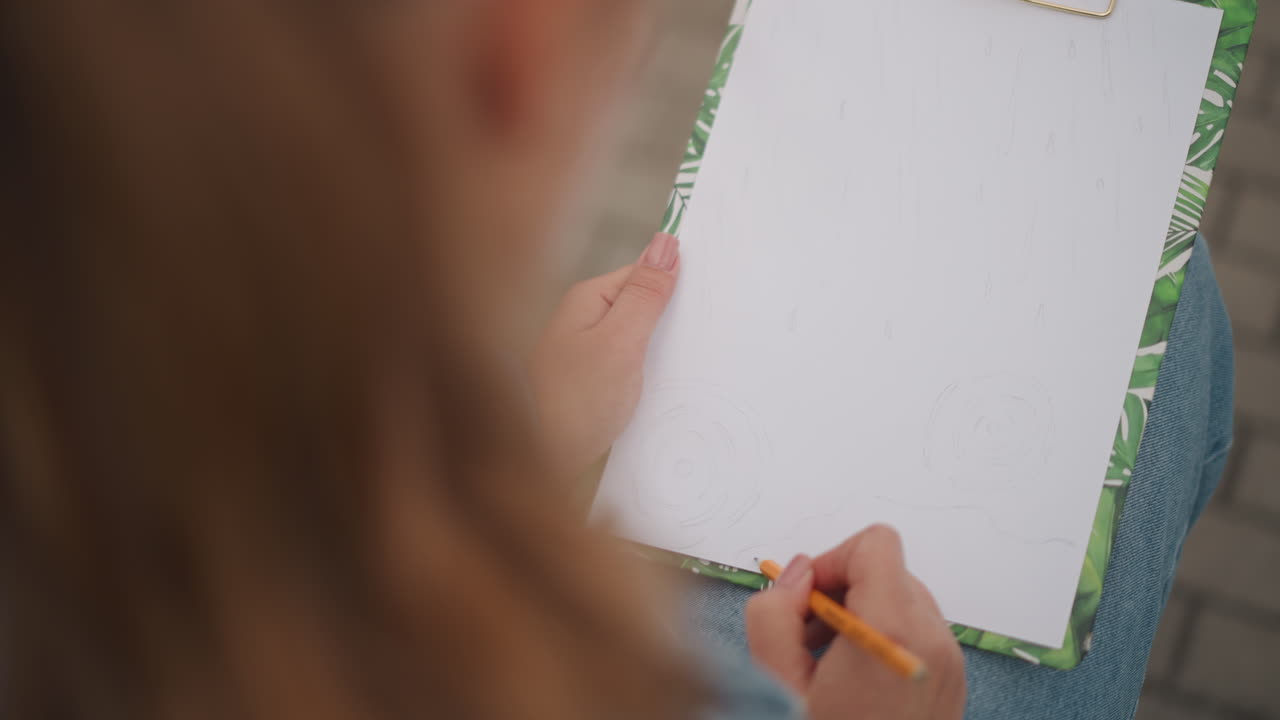Studente universitario che prende appunti su un blocco vicino al marciapiede, con giacca di jeans visibile, matita sospesa su carta ruvida, movimento fermo della mano, atmosfera da campus urbano, scena di studio pensierosa, scatto spontaneo.
