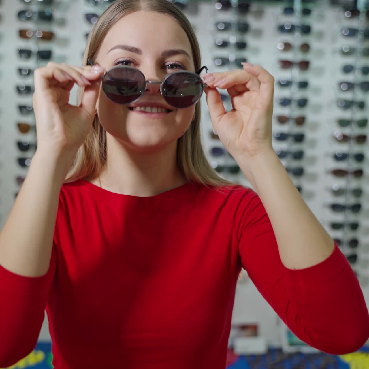 Portrait of smiling girl in black sunglasses. Attractive young woman is trying on stylish eyeglasses on the background of new spectacles at optical store.