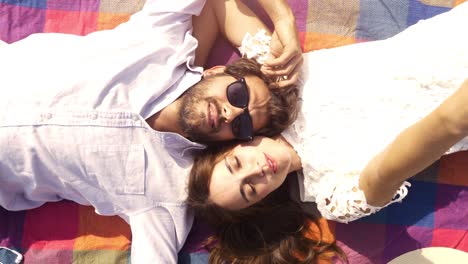 feliz y encantadora pareja joven acostada en una manta de colores en el parque mirando al cielo apuntando a las nubes romántico con gafas de sol de guitarra hermosa chica atractiva vista superior cámara giratoria en cámara lenta