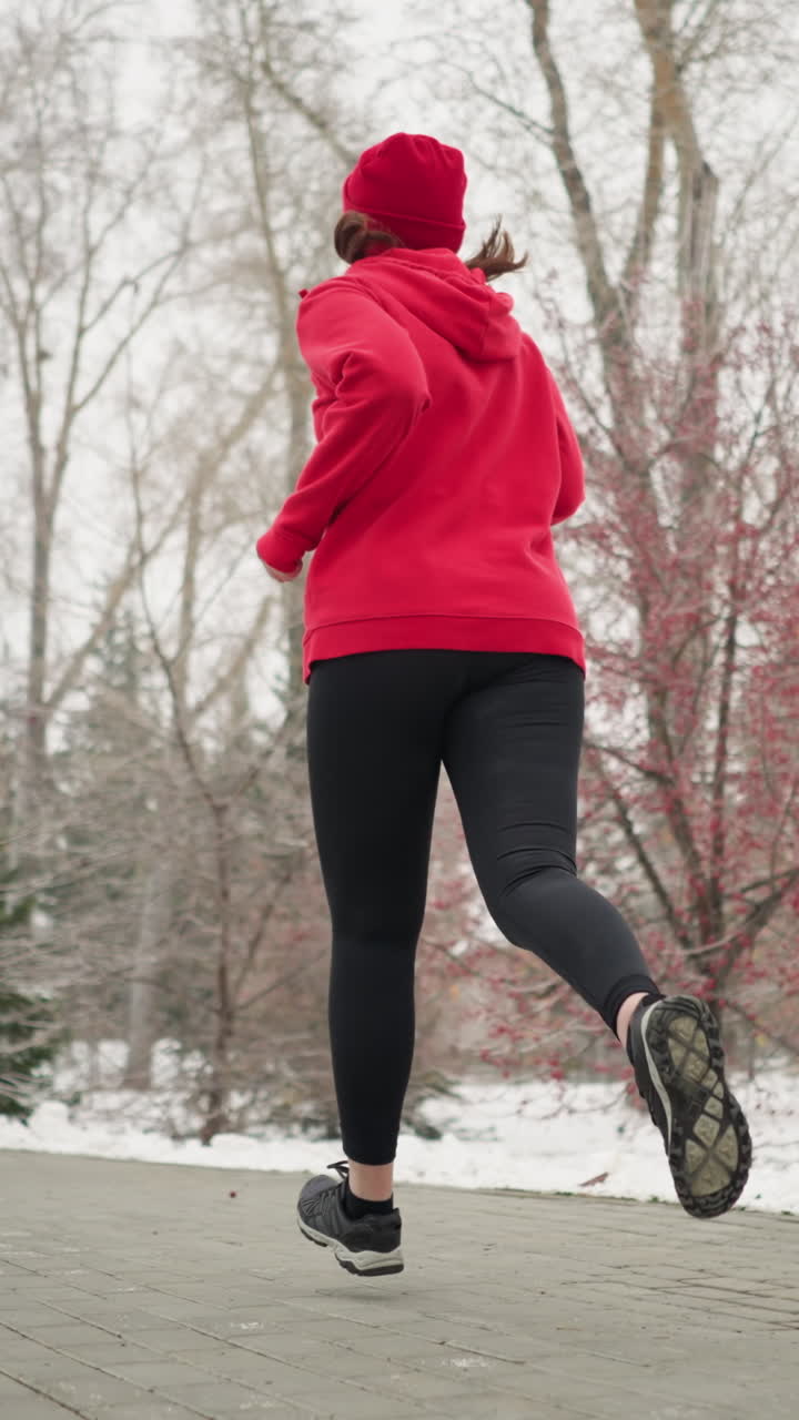back view middle aged woman in red hoodie and beanie jogging along snowy park pathway flanked by frosty trees and pines under serene foggy winter sky conveying calm endurance outdoors