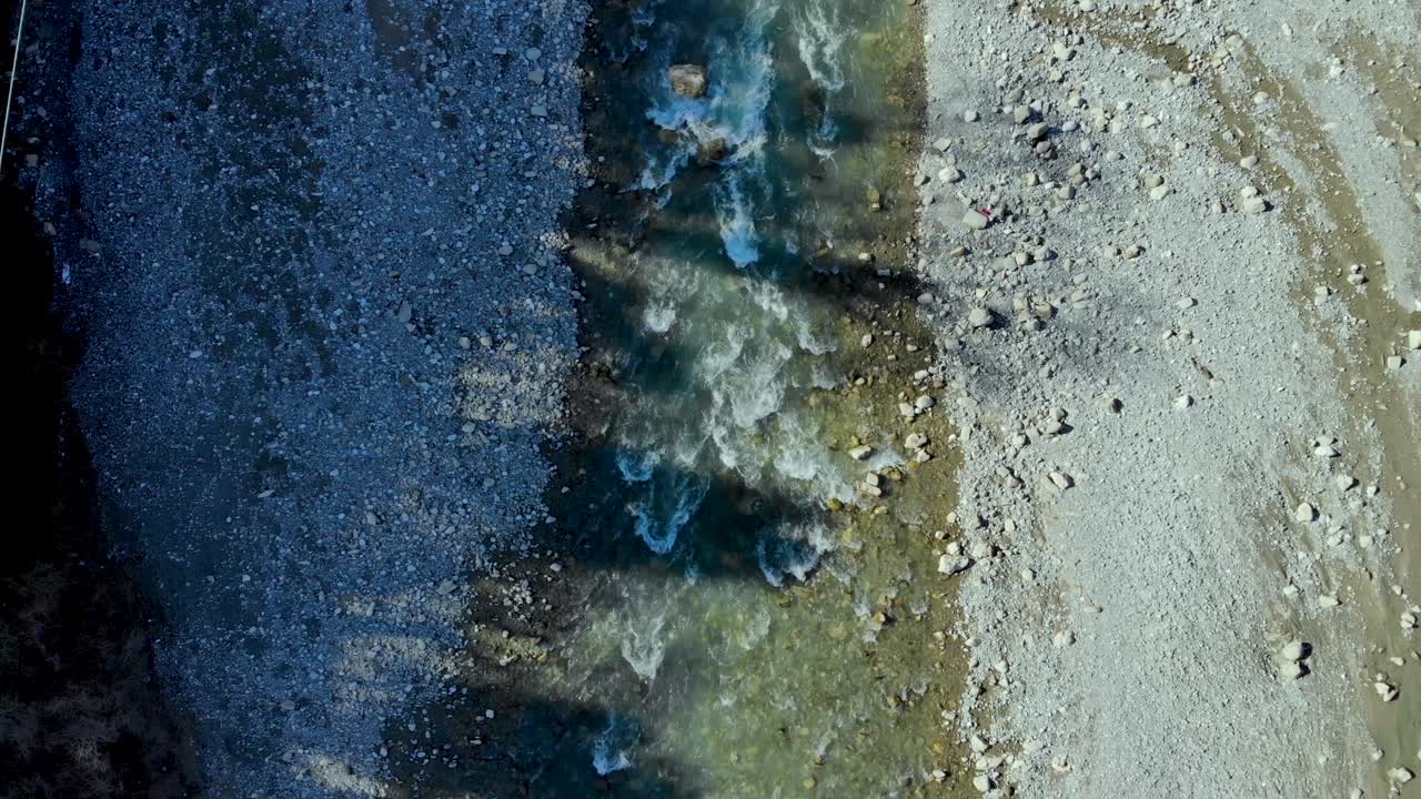 River streaming across pebbles and rocks splashing and foaming, shadow of trees over water surface, top down aerial view