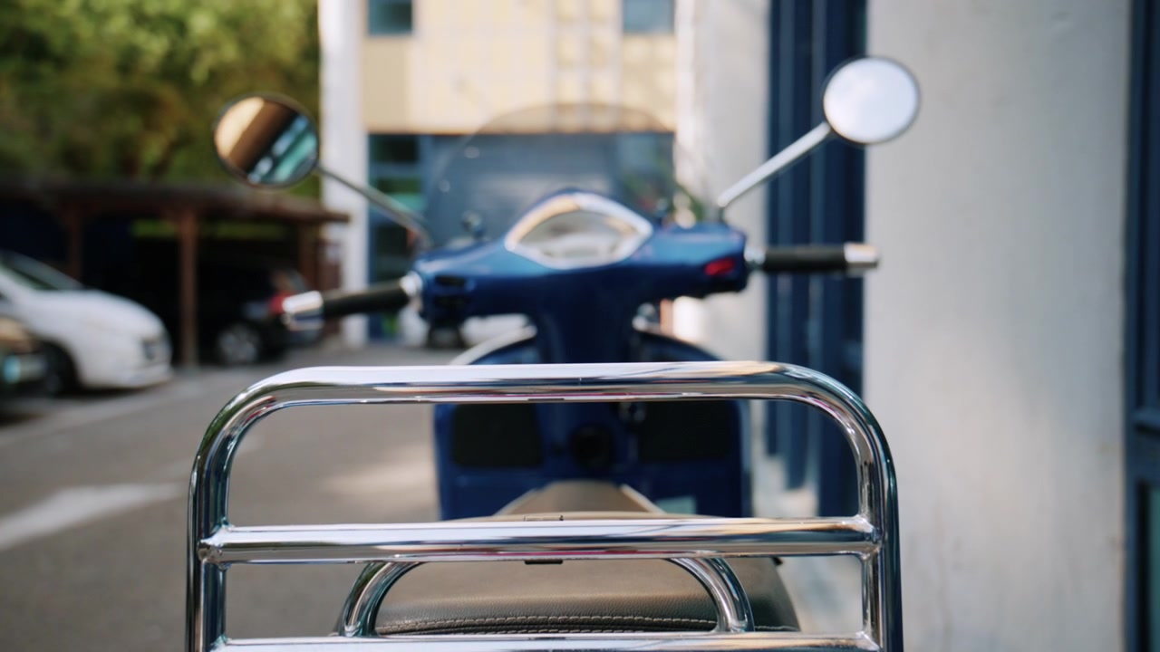 Rear view of a parked blue scooter near a building entrance, with chrome rack and mirrors in soft focus