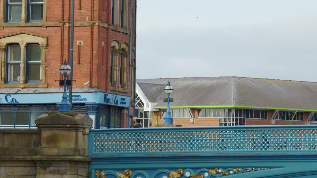 Pedestrians cross a blue metal bridge beside historic brick building under cloudy daylight, static shot